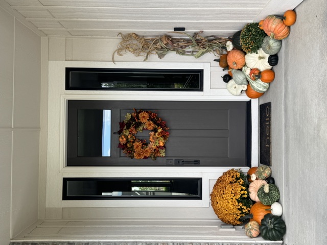 Front porch with a fall wreath on a dark door, pumpkin and gourd arrangements on both sides, cornstalks, and a potted golden mum